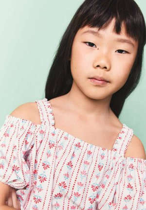 Blouse rose clair à épaules dénudées avec un motif floral rouge et bleu, manches à volants et fines bretelles sur un fond vert.