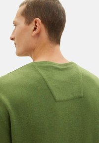 Grön stickad tröja med en fyrkantig detalj på baksidan av halsringningen, tillverkad av mjukt material med en slät textur och en tajt passform.