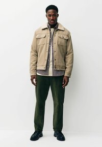 Beige suede jacket with a shearling collar, buttoned front, two chest pockets; layered over a checkered shirt and olive green pants. Black shoes.