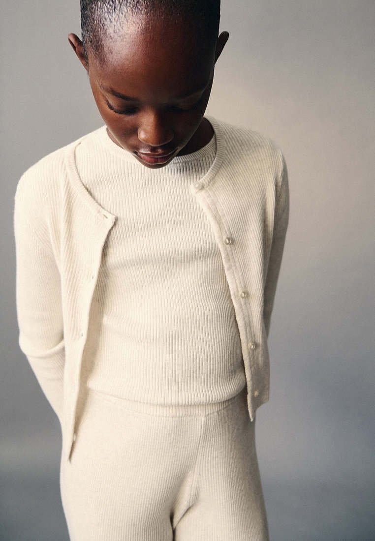 Jeune personne aux cheveux courts portant un pull côtelé crème et un pantalon, regardant vers le bas avec les mains dans le dos sur un fond neutre.