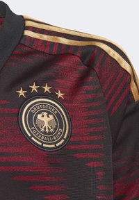 Musta ja punainen urheiluverkkopaita, jossa on rakenteellinen muotoilu, kultaiset yksityiskohdat sekä Saksan jalkapalloliiton logo, jonka yläpuolella on neljä kultatähteä.