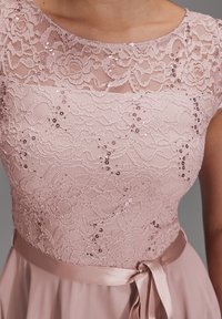 Čipkana haljina u mekoj rozoj boji s uzorkom cvjetova od šljokica. Ima satenasti pojas i mašnu, glatku tkaninu suknje. Elegantan dizajn dekoltea.