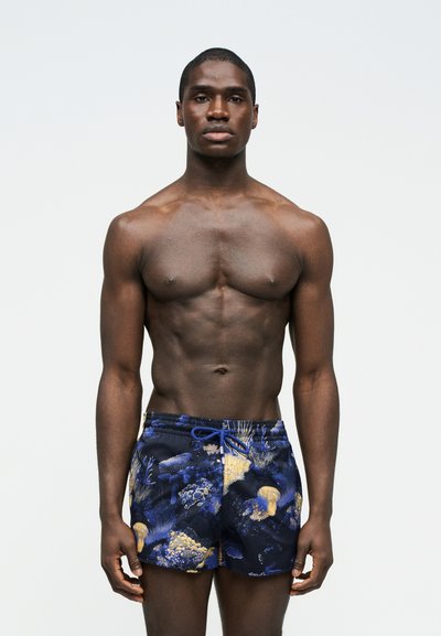 Άνδρας χωρίς φανέλα με μυώδη σωματότυπο, που φορά μπλε μαγιό με σχέδια υποβρύχιων κοραλλιών και μέδουσες, στέκεται μπροστά σε απλό φόντο.