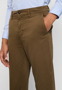 Bruna chinos med en slät textur, som har en framficka, subtila sömmar och en knäppning. Parat med en blårandig skjortärm.