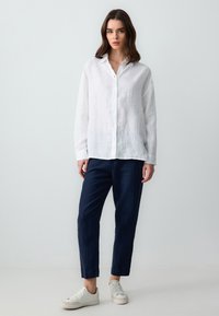 Chemise blanche en lin à boutons avec une coupe décontractée, associée à un pantalon en lin bleu marine et des baskets blanches. Le tissu semble léger et texturé.