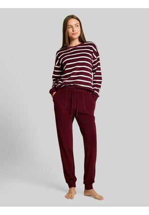 Vrouw staat blootsvoets in een bordeaux gestreepte top met lange mouwen en bijpassende broek met aantrekkoord tegen een effen grijze achtergrond.
