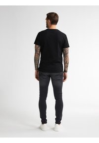 Camiseta negra de manga corta con cuello redondo, combinada con jeans ajustados gris oscuro y zapatillas blancas, vista desde detrás.