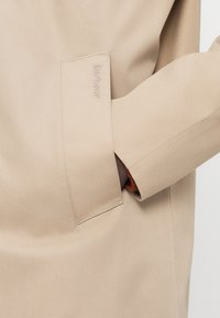 Μπεζ παλτό με λεία υφή, που διαθέτει ορατή τσέπη και λογότυπο Barbour. Το μανίκι έχει καθαρό φινίρισμα με μοντέρνα μορφή.