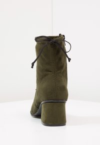 Botín de ante verde oliva hasta el tobillo con un tacón de bloque, que presenta un detalle de cordones y cierre de cremallera. Textura suave con costuras mínimas.