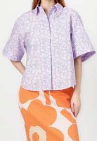 Chemise lavande à manches courtes avec un imprimé floral intégral, col boutonné et coupe décontractée. Associée à une jupe orange à motif graphique.