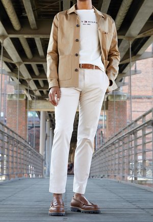 Tan button-up jas over een wit T-shirt, gecombineerd met crèmekleurige broek en bruine loafers. Kenmerkt zich door een gestructureerde stof en zichtbare stiksels.