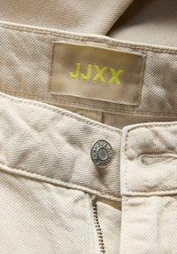 Tissu en denim beige avec une étiquette cousue affichant "JJXX" en jaune vif, un bouton-pression argenté et un détail d'ouverture éclair.