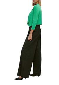 Mujer vistiendo un blazer corto de color verde brillante, pantalones anchos negros y tacones de punta negra, de pie de perfil.