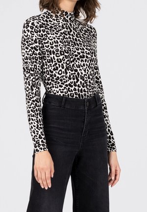 Femme portant un col roulé à manches longues imprimé léopard noir et blanc, rentré dans un jean noir taille haute, sur un fond uni.