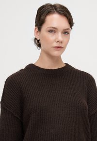 Tmavě hnědý pletený svetr s ribovanou texturou, kulatým výstřihem a mírně oversized střihem, s volnějším ramenem a minimálním zdobením.