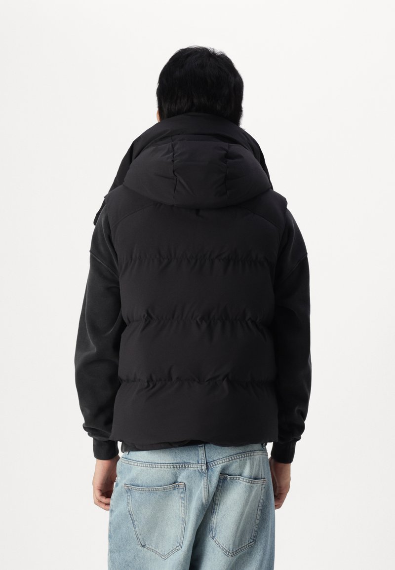 Chaqueta de plumas negra con capucha, que presenta un diseño acolchado y puños acanalados. Llevada sobre una camiseta de manga larga gris y jeans de denim claros.