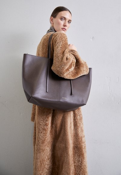Large, textured dark brown tote bag with two long handles. Bag features a minimalist design and topstitching for added detail.
