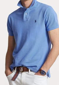 Lichtblauwe poloshirt met een gestructureerde kraag, twee knopen en een klein logo. Gerd gedragen met witte broek en een multicolore gestreepte riem.