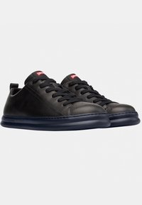 Le sneakers in pelle nera presentano punte arrotondate, lacci piatti e una suola in gomma blu navy con texture. Sono dotate di una piccola etichetta con logo rosso sulla linguetta.