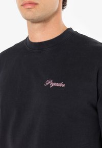Tricou din bumbac negru, cu guler rotund și mâneci scurte. Dispune de un text brodat roz care spune "Pegador" pe piept.