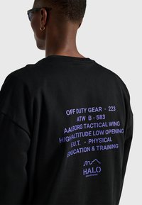 Czarna, oversize'owa koszulka z fioletowym napisem na plecach, zawierającym różne frazy oraz logo na dole. Wykonana z miękkiej bawełny o luźnym kroju.