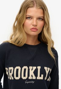 Marine sweatshirt met een gestructureerde afwerking, met "BROOKLYN" in grote beige letters en "Superdry" in een kleinere script eronder.
