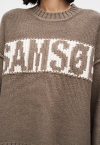Καφέ πλεκτό πουλόβερ με λευκό μπλοκ σχέδιο και το κείμενο "SAMSON." Η λαιμόκοψη έχει ριμπ ύφανση και το ύφασμα έχει υφή πλέξης.