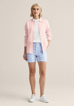 Kvinde iført lyserød skjorte med knapper over en hvid tekstureret top, blå blomstrede shorts og hvide sneakers, stående med den ene hånd i lommen.