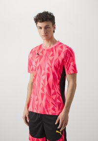 Camiseta deportiva rosa con un patrón geométrico, de manga corta, cuello redondo y acentos laterales en negro; combinada con pantalones cortos negros que tienen un logo amarillo.