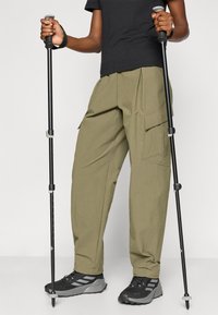 Παντελόνια cargo σε ελιά πράσινο με πιέτες, πλαϊνές τσέπες και άνετη εφαρμογή, συνδυασμένα με μαύρα παπούτσια πεζοπορίας και μπαστούνια trekking.
