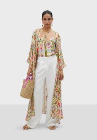 Un kimono fluido e stampato in colori pastello sopra un top colorato, abbinato a pantaloni a gamba larga bianchi e a una borsa intrecciata con accenti rosa.