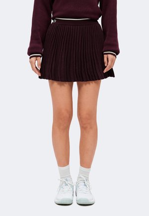 Donna che indossa una gonna plissettata scura e un maglione abbinato, calzini bianchi alla caviglia e scarpe sportive bianche, in piedi su uno sfondo chiaro.