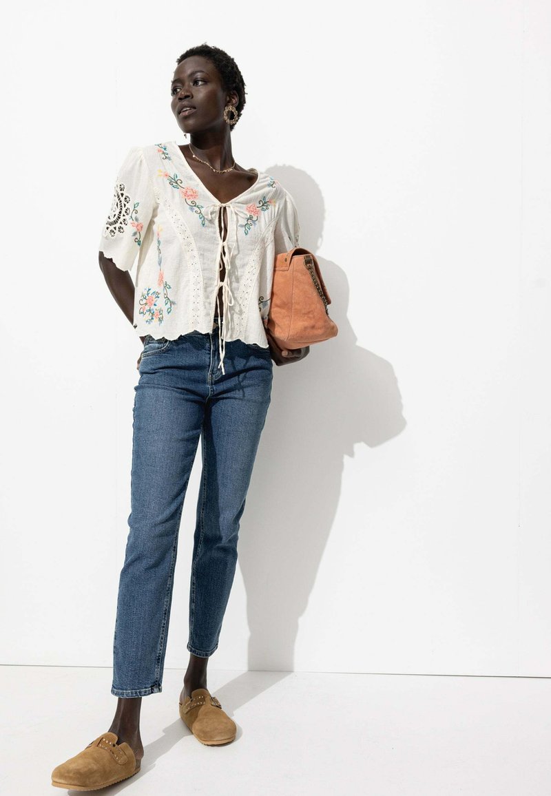 Blouse blanche brodée à manches courtes, jean bleu clair et chaussures slip-on marron. Portant un sac à main beige avec une bandoulière en accent.