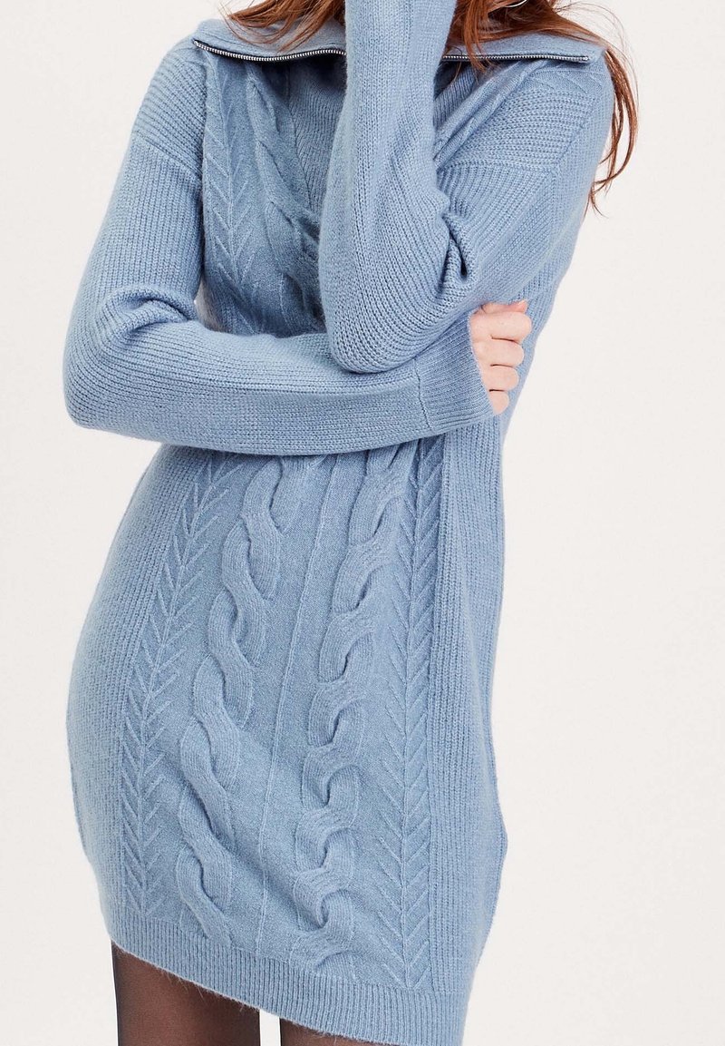 Femme portant une robe-pull tricotée bleu clair à torsades, manches longues et col zippé, bras croisés sur la poitrine.