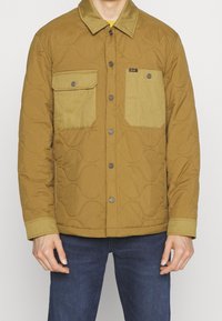 Gesteppte braune Jacke mit einem helleren beigen Kragen und Taschen, ausgestattet mit Druckknöpfen und einem geometrischen Muster auf dem Stoff.
