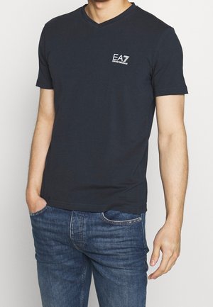 Mørkeblå bomulds t-shirt med v-udskæring og korte ærmer, med en lille hvid EA7-logo på venstre bryst. Båret med cowboybukser.