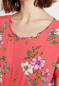 Koralbluse mit rundem Ausschnitt, die mit einem floralen Muster aus pinken, weißen und gelben Blumen verziert ist, akzentuiert durch grüne Blätter. Weicher Stoff.
