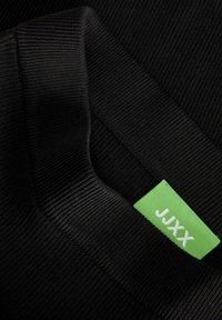 Tessuto nero a coste con un primo piano di un colletto e un'etichetta verde con il testo "JJXX." La texture è liscia e lavorata a maglia.