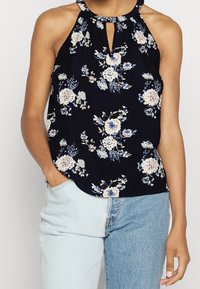 Haut halter fleuri en navy avec des fleurs bleu clair et crème. Présente un col en forme de trou de serrure et un tissu doux et léger.