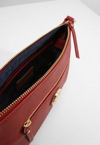 Czerwony skórzany portfel na nadgarstek z fakturą, z złotym zamkiem i zapięciem, ukazujący czarną tkaninę wewnętrzną z małą brązową metką.