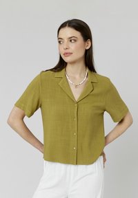 Blouse à manches courtes, de couleur olive, avec un devant à boutons, un col et un tissu texturé. Portée avec un pantalon clair et un collier de perles.