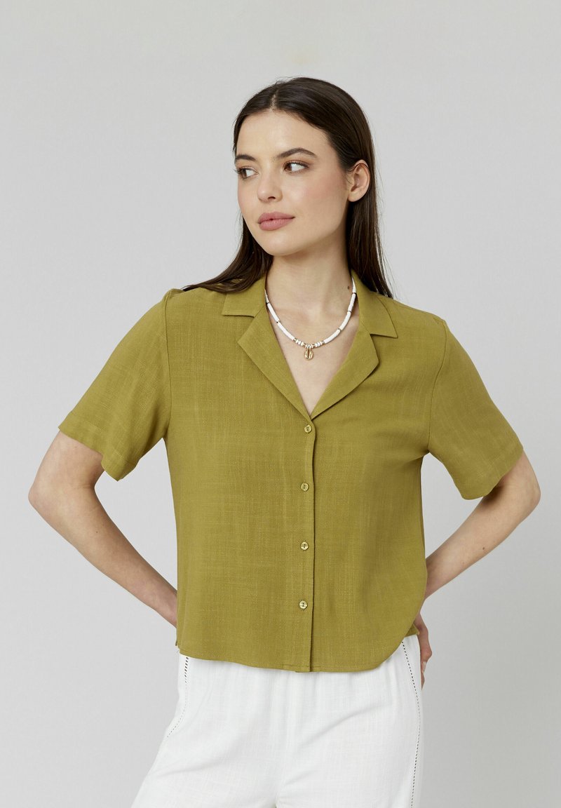 Blouse à manches courtes, de couleur olive, avec un devant à boutons, un col et un tissu texturé. Portée avec un pantalon clair et un collier de perles.
