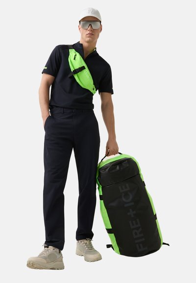 Černý batoh s neonově zelenými prvky a textem "FIRE + ICE", který je kombinován s odpovídající ledvinkou. Model má na sobě námořnický polo a kalhoty, světlé tenisky.