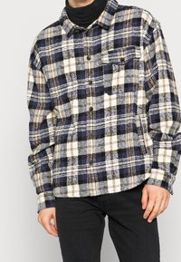 Rutigt flanellskjorta i marinblått, krämigt och beige. Har knappstängning i fronten, krage och en enskild bröstficka. Mjuk textur, långa ärmar.