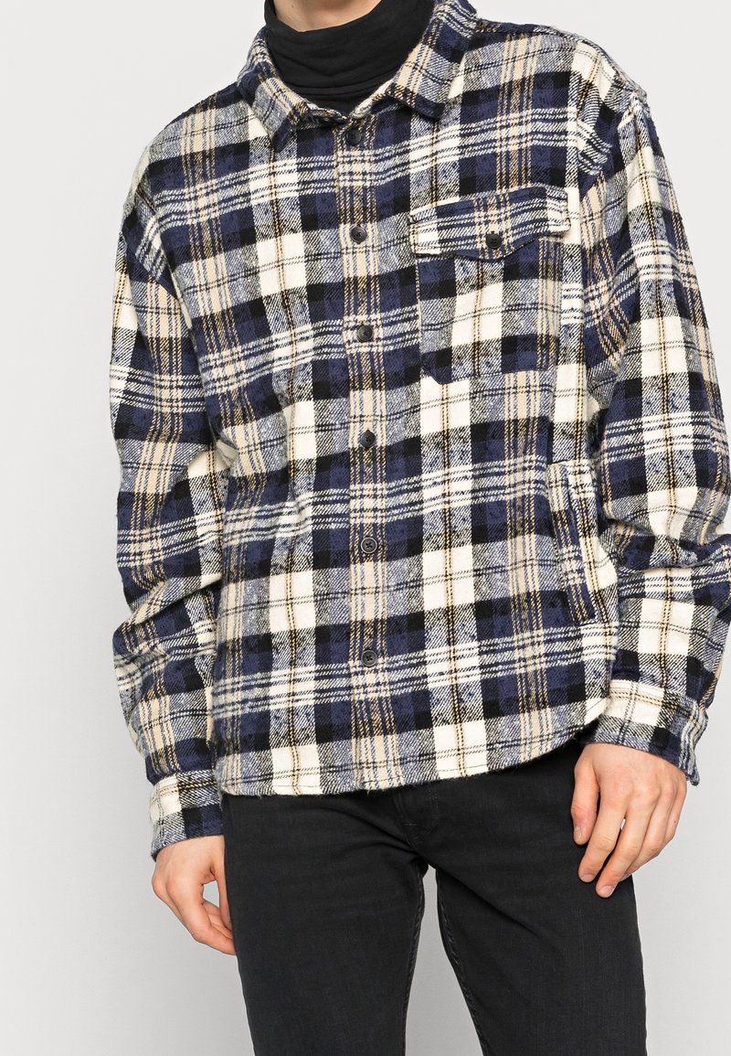 Rutigt flanellskjorta i marinblått, krämigt och beige. Har knappstängning i fronten, krage och en enskild bröstficka. Mjuk textur, långa ärmar.