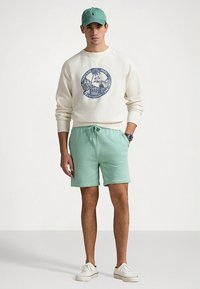 Sweatshirt creme claro com gráfico redondo, calções com cordão teal e boné a condizer. Tênis brancos completam o visual casual.
