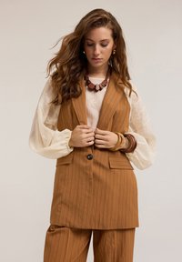 Chaleco marrón a rayas con bolsillos frontales, combinado con una blusa crema con detalles bordados. Los accesorios incluyen un collar de cuentas de madera.