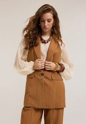 Gilet marrone a righe con tasche frontali, abbinato a una blusa crema con dettagli ricamati. Gli accessori includono una collana di perline in legno.