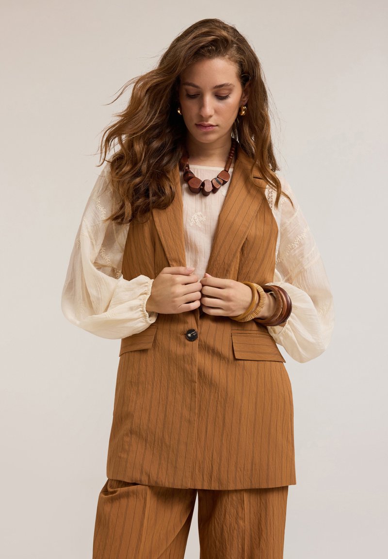 Chaleco marrón a rayas con bolsillos frontales, combinado con una blusa crema con detalles bordados. Los accesorios incluyen un collar de cuentas de madera.