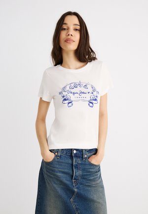 Νεαρή γυναίκα με μεσαίου μήκους σκούρα μαλλιά που φοράει ένα λευκό μπλουζάκι με φλοράλ σχέδιο και μια μπλε τζιν φούστα με τα χέρια στις τσέπες.